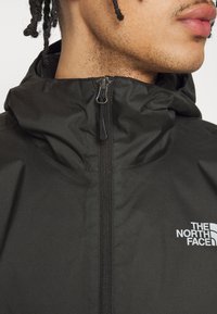 Black waterproof jacket with a zipped front, adjustable hood, and embossed branding on the chest. Smooth texture and minimalistic design.
