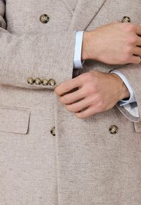 Muž si upravuje manžetu béžového štruktúrovaného blejzra s hnedými gombíkmi cez rukávy svetlomodrej košele.