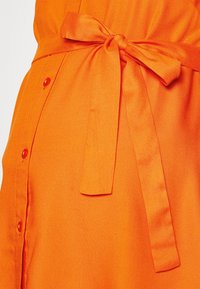 Vestido laranja com um cinto atado na cintura, apresentando botões ao longo do lado. A textura suave e o design ajustado realçam a silhueta.