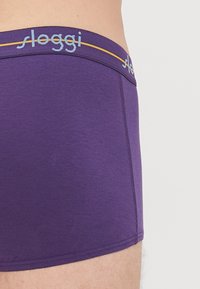 Nahaufnahme einer Person in lila Boxershorts mit einem markierten lila Bund, auf dem der Text "sloggi" und ein dünner gelber Streifen zu sehen sind.