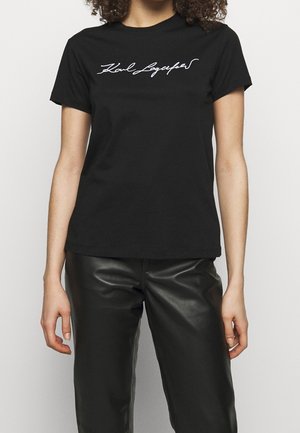 Kobieta w czarnym dopasowanym T-shircie z białym napisem „Karl Lagerfeld” oraz w czarnych spodniach z ekologicznej skóry, stojąca na jednolitym tle.