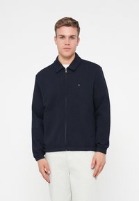 Hajózási kék, zipzáros dzseki, galléros nyakkal, hosszú ujjakkal és oldalsó zsebekkel. Kis hímzett logó található a mellkason.