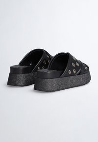 Un par de sandalias de plataforma negras con suelas gruesas brillantes y una correa ancha con un diseño sobre un fondo claro.