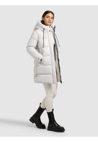 Žena u dugoj bijeloj puhanoj jakni s kapuljačom, bijelim helankama i crnim masivnim čizmama, stoji s rukama u džepovima na bijeloj pozadini.