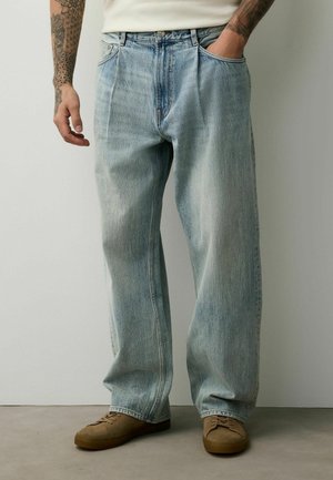 Man with tattooed arms wearing loose, light blue jeans and brown suede sneakers, with one hand in pocket, standing against plain wall.