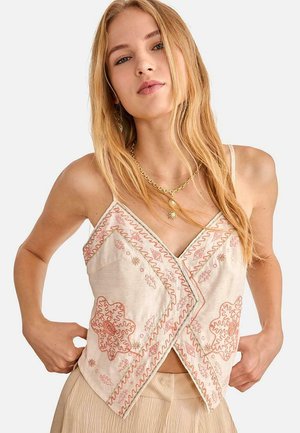 Joven con cabello rubio largo que lleva una blusa sin mangas beige bordada y un collar dorado sobre un fondo liso.