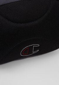 Gros plan sur un tissu texturé noir avec un petit emblème cousu présentant un contour blanc et un détail intérieur rouge sur une surface courbée.