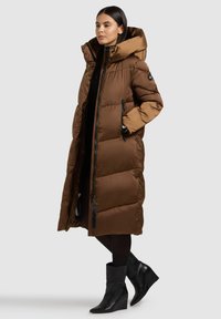 Dolga, oblazinjena rjava jakna z veliko kapuco in žepi z zadrgami. Ima sijočo teksturo in črne tehnične dodatke. Oblečena preko črnega oblačila.