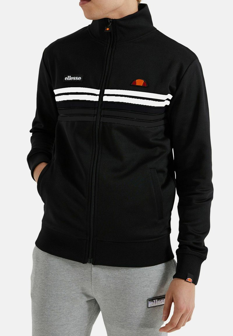 Man in een zwarte ritsjack met witte strepen en logo's, gecombineerd met grijze broek met een logopatch op de dij.