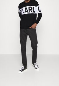 Musta neule valkoisella "KARL" -logokujalla, yhdistetty tummanharmaisiin housuihin ja mustiin tennareihin. Neuleessa on pyöreä kaula-aukko ja resorit hihansuissa.