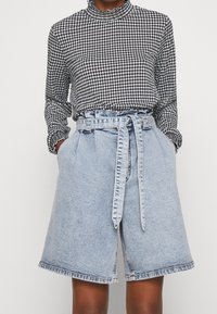 Osoba oblečená v černobílé houndstooth dlouhém rukávu, zasunutém do světle modrých šortek s vysokým pasem, které mají odpovídající pásek.