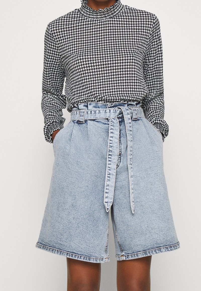 Osoba oblečená v černobílé houndstooth dlouhém rukávu, zasunutém do světle modrých šortek s vysokým pasem, které mají odpovídající pásek.