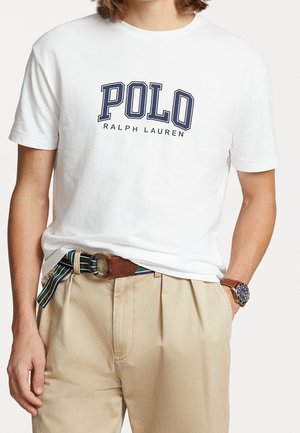 Osoba nosząca białą koszulkę Polo Ralph Lauren, beżowe spodnie z paskiem w paski oraz zegarek na lewym nadgarstku.