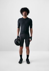 Maglia da ciclismo nera a maniche corte con chiusura a zip, abbinata a pantaloni corti neri. Il modello tiene un casco nero e indossa scarpe da ciclismo e guanti.