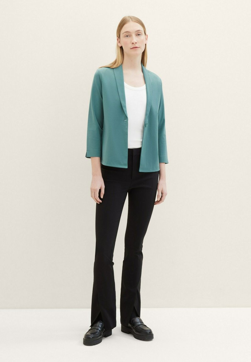 Teal blazer met 3/4 mouwen en een sluiting met één knoop, gecombineerd met slim-fit zwarte broek met splitten aan de zoom, gedragen over een witte top.