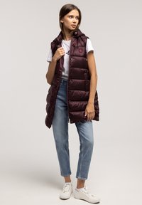 Burgundowy kamizelka puchowa z kapturem, charakteryzująca się pikowanym wzorem i napami, w zestawie z białą koszulką i niebieskimi dżinsami.