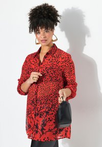 Blusa roja con patrón de puntos negros y diseño floral, de manga larga, con botones en el frente, combinada con un pequeño bolso negro.