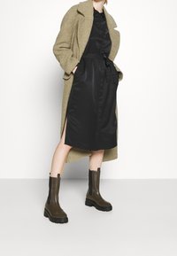 Cappotto texturizzato verde oliva su un abito satinato nero con vita legata, abbinato a stivaletti alla caviglia marrone scuro con suole in gomma spessa.