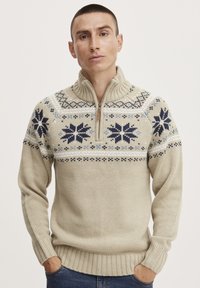 Beige neulepullover, jossa on korkea kaulus. Rinnassa on sinisiä ja tummansinisiä lumihiutaleita sekä ribbikujetteet hihansuissa ja helmassa.