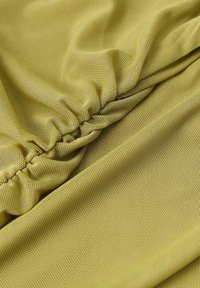 Zbliżenie na żółto-zielony, teksturowany materiał z marszczonym elastycznym szwem, tworzącym fałdy i głębię.