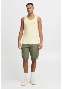 Heled kollane tank top, millel on väike graafikuga tasku ja mis on kombineeritud oliivroheliste cargo-lühkpükstega ning valgete tennisekatega, millel on tekstuuriga tallad.