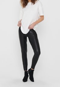 Svarta läderliknande leggings ihop med en lös vit t-shirt; modellen bär svarta ankelstövlar; slät textur; figurnära design.