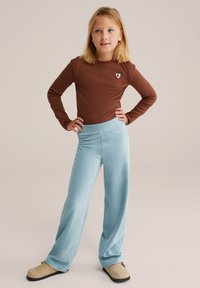 Camiseta marrón de manga larga con un parche en forma de corazón, combinada con pantalones de pierna ancha de canalé en azul claro. El conjunto se completa con zapatos beige tipo slip-on.