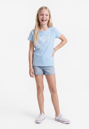 Smilende blond pige stående med hånden på hoften iført lyseblå t-shirt med hjerte-logo, cowboyshorts og hvide sneakers på hvid baggrund.