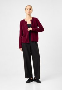 Burgundský cardigan vyrobený z měkké tkaniny, s kulatým výstřihem, zapínáním na knoflíky a dvěma předními kapsami, kombinovaný s černými kalhotami s širokými nohavicemi.