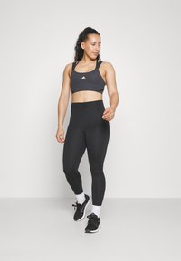 Čierny športový crop top s tenkými ramienkami a logom, skombinovaný s čiernymi legínami s vysokým pásom. Žena má na sebe čierne tenisky s bielymi akcentmi.