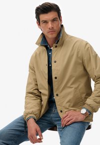 Beige, waterdichte jas met een hoge kraag, drukknoopsluitingen en elastische manchetten over een denim shirt, gecombineerd met blauwe jeans en een horloge.