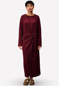 Femme aux cheveux courts et bouclés portant une longue robe bordeaux avec un nœud sur le côté, des chaussettes beiges et des sandales fermées à bout léopard, se tenant devant un fond blanc.