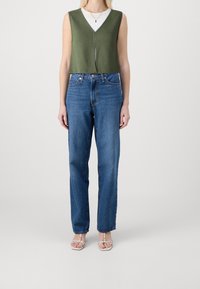 Zielony bezrękawnik dzianinowy z dekoltem w serek, nałożony na biały T-shirt z okrągłym dekoltem. W połączeniu z niebieskimi dżinsami z wysokim stanem i białymi sandałami.