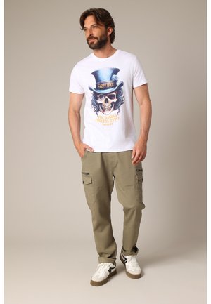 Man staand, draagt een wit t-shirt met schedel in hoge hoed afbeelding en kaki cargobroek met witte sneakers op een neutrale achtergrond.