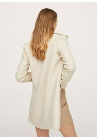 Mujer con cabello largo de color castaño claro, vistiendo un abrigo beige y una falda marrón, de pie con las manos en las caderas, de espaldas contra un fondo liso.