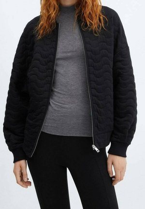 Kobieta z czerwonymi włosami, ubrana w czarną pikowaną kurtkę bomberkę na szarym golfie i czarnych spodniach, stojąca na prostym tle.