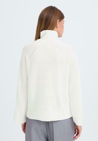 Vit stickad tröja med hög krage, ribbad textur och sänkt axel, tillsammans med grå byxor. Sett bakifrån.