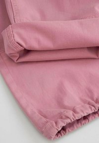 Jasnoróżowa tkanina o gładkiej fakturze, z podwiniętym wykończeniem i marszczonym elastycznym detalem na dolnej krawędzi. Widoczne szwy.