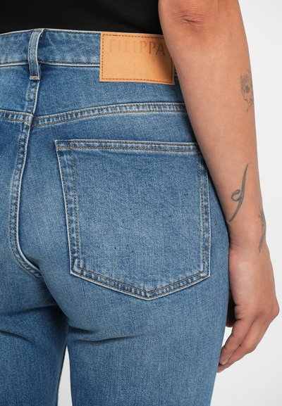 Jeans de mezclilla azul con talle alto, que presentan una etiqueta de cuero marrón con el logo "Filippa K" en relieve y bolsillos traseros con costuras en contraste.