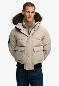 Beżowa kurtka puchowa z kapturem wykończonym futrem, czarnymi akcentami i dwiema przednimi kieszeniami. Noszona na szarym rozpinanym swetrze i koszuli w kratę. Ciemne dżinsy.