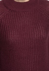 Pull en maille bordeaux avec une texture à côtes, doté d'un col rond et de manches raglan. Le matériau semble doux et chaud.