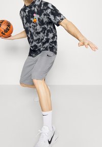 Persona driblando un balón de baloncesto naranja, vistiendo una camiseta de tirantes negra y gris con diseño de tie-dye, pantalones cortos grises, calcetines blancos de Nike y zapatillas blancas de Nike.