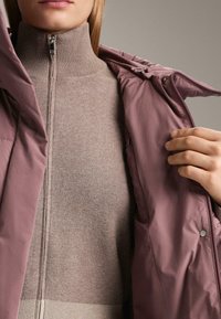 Jasnofioletowa kurtka zewnętrzna z wysokim kołnierzem, rozpięta, odsłaniająca beżową, dzianą warstwę wewnętrzną. Z widocznym zamkiem błyskawicznym i zatrzaskami.