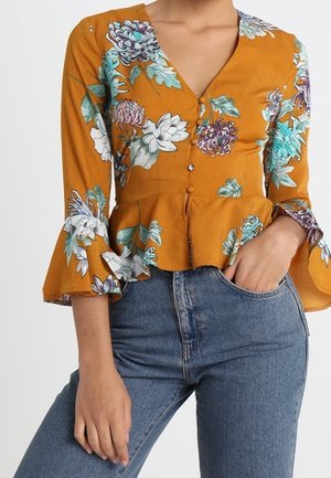 Kvinde iført sennepsgul bluse med blomsterprint, V-udskæring, knapper foran, klokkeærmer og peplumkant, kombineret med blå jeans.