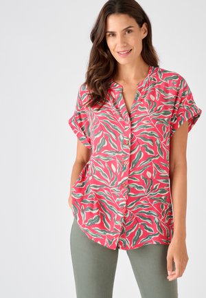 Mujer con camiseta roja de manga corta con estampado de hojas verdes y blancas, combinada con pantalones verde oliva, de pie con las manos en los bolsillos.