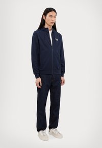 Marineblå zip-up hettegenser med en teksturert overflate, med en frontlomme og logo, kombinert med mørke denimjeans og lyse joggesko.