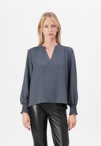 Grå langermet bluse med V-hals, med smockede mansjetter og løs passform. Glatt tekstur og lett materiale. Kombinert med svarte bukser.