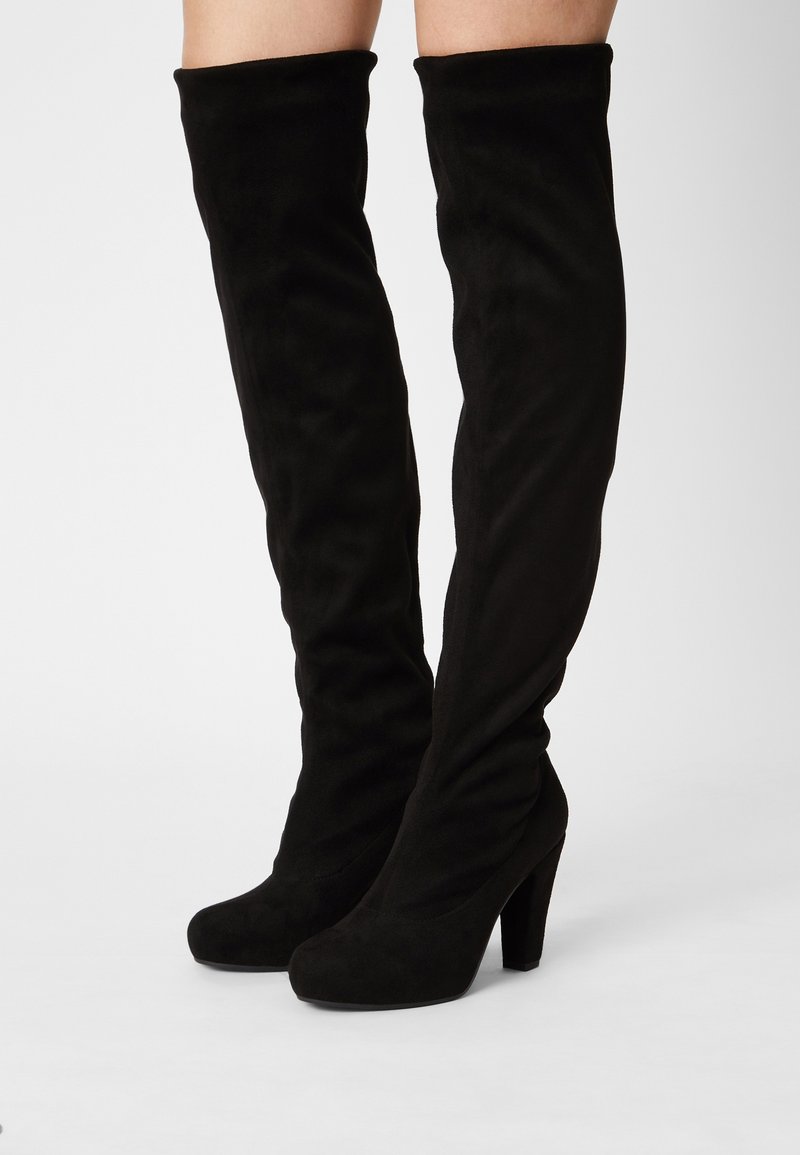 Bottes hautes en daim noir jusqu'à la cuisse avec des bouts arrondis et des talons blocs hauts portées sur des jambes nues.
