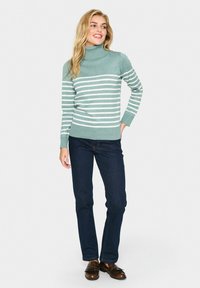 Svītrainais augstais kakls džemperis gaismas teal un baltā krāsā, izgatavots no mīkstas trikotāžas. Pāris ar tumši ziliem džinsiem un brūniem mokasīniem.