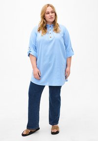 Hellblaues Tunika-Shirt mit Kragen, Knopfdetails und hochgerollten Ärmeln, kombiniert mit dunkelblauen Jeans und braunen Sandalen. Schlichtes Design.